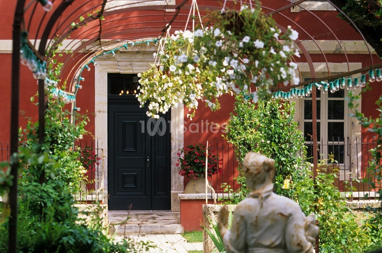 Entrée d'une maison rouge avec porte noire, encadrée par des fleurs suspendues et un jardin verdoyant au premier plan.