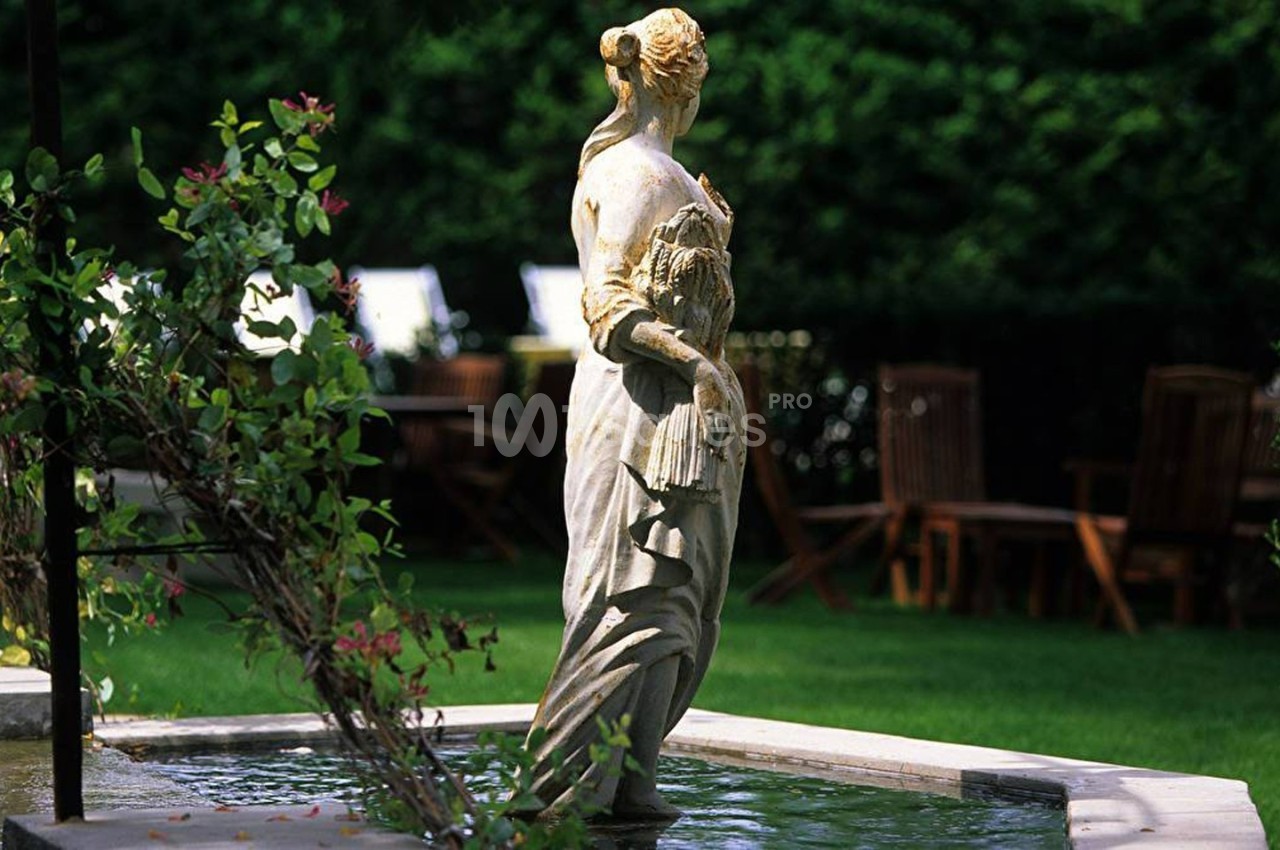 Statue en pierre représentant une femme tenant des épis de blé, placée au centre d'un bassin dans un jardin verdoyant.
