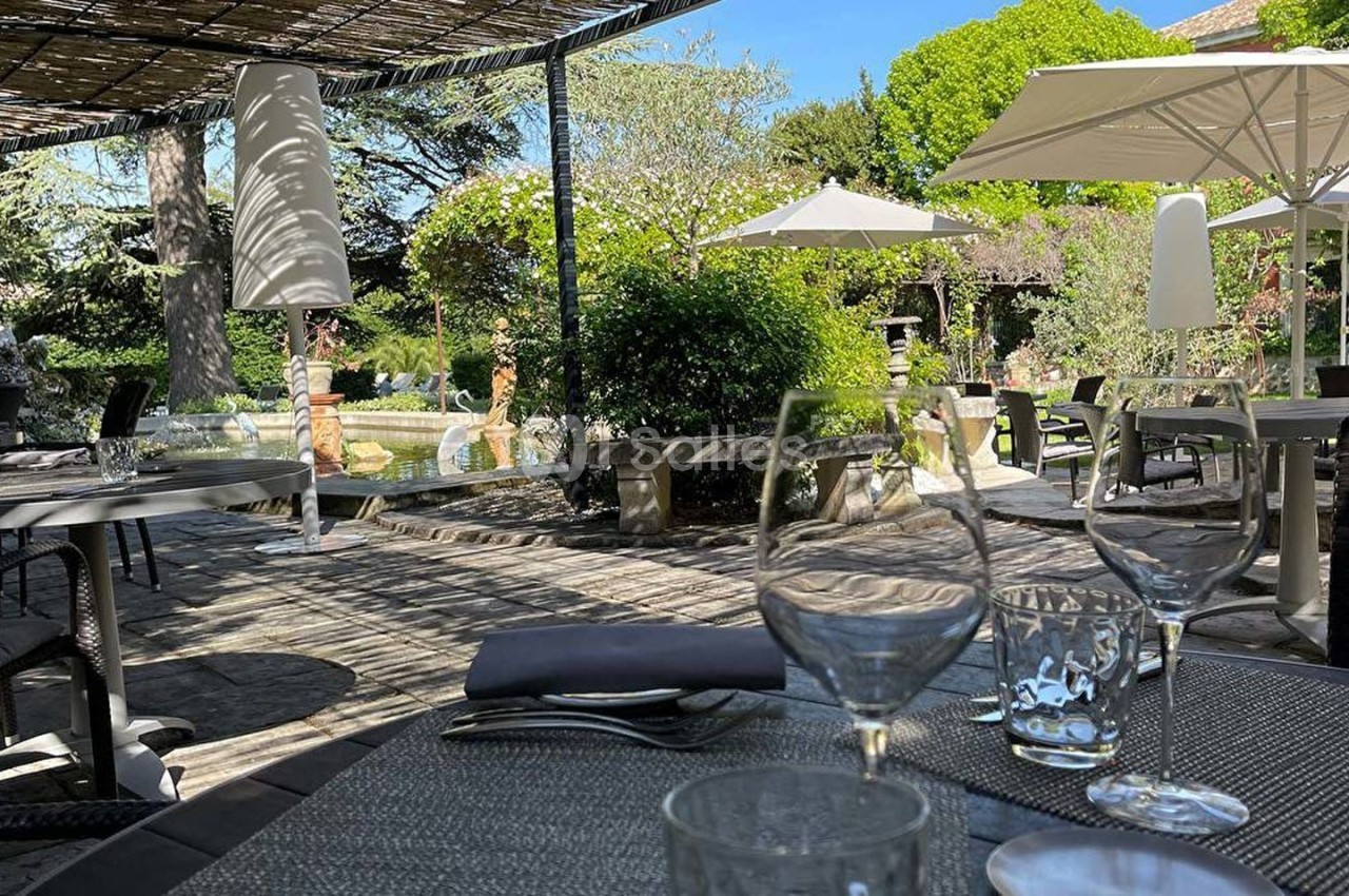 Terrasse ombragée avec tables dressées, vue sur un jardin verdoyant et parasols blancs par une journée ensoleillée.