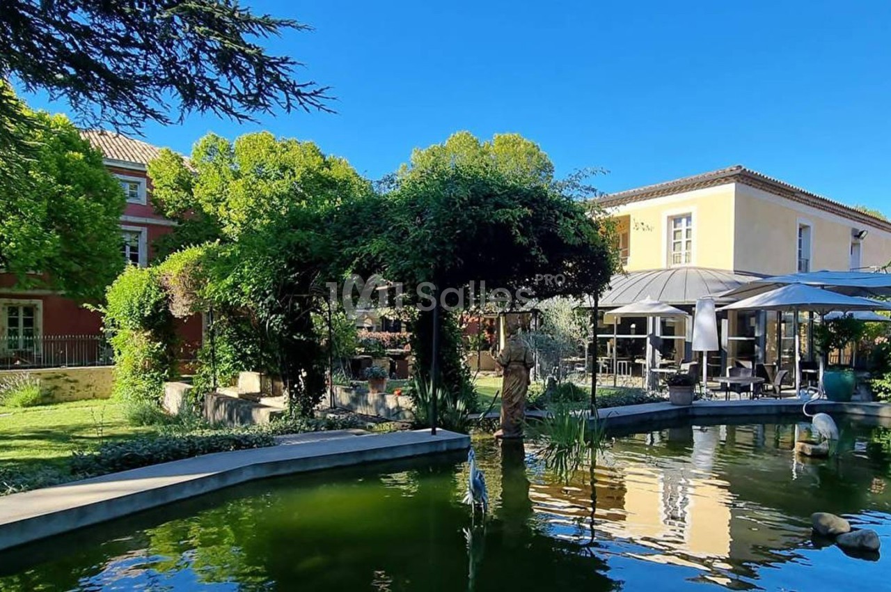 Jardin avec bassin, végétation luxuriante, terrasse ombragée et bâtiments en arrière-plan sous un ciel bleu.
