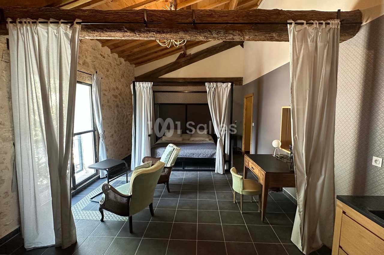 Chambre avec lit à baldaquin, fauteuils, bureau en bois, grandes fenêtres et poutres apparentes sous un plafond en bois.
