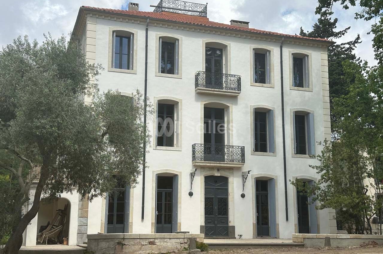 Façade d'une grande maison blanche à trois étages avec balcons en fer forgé, entourée d'arbres.