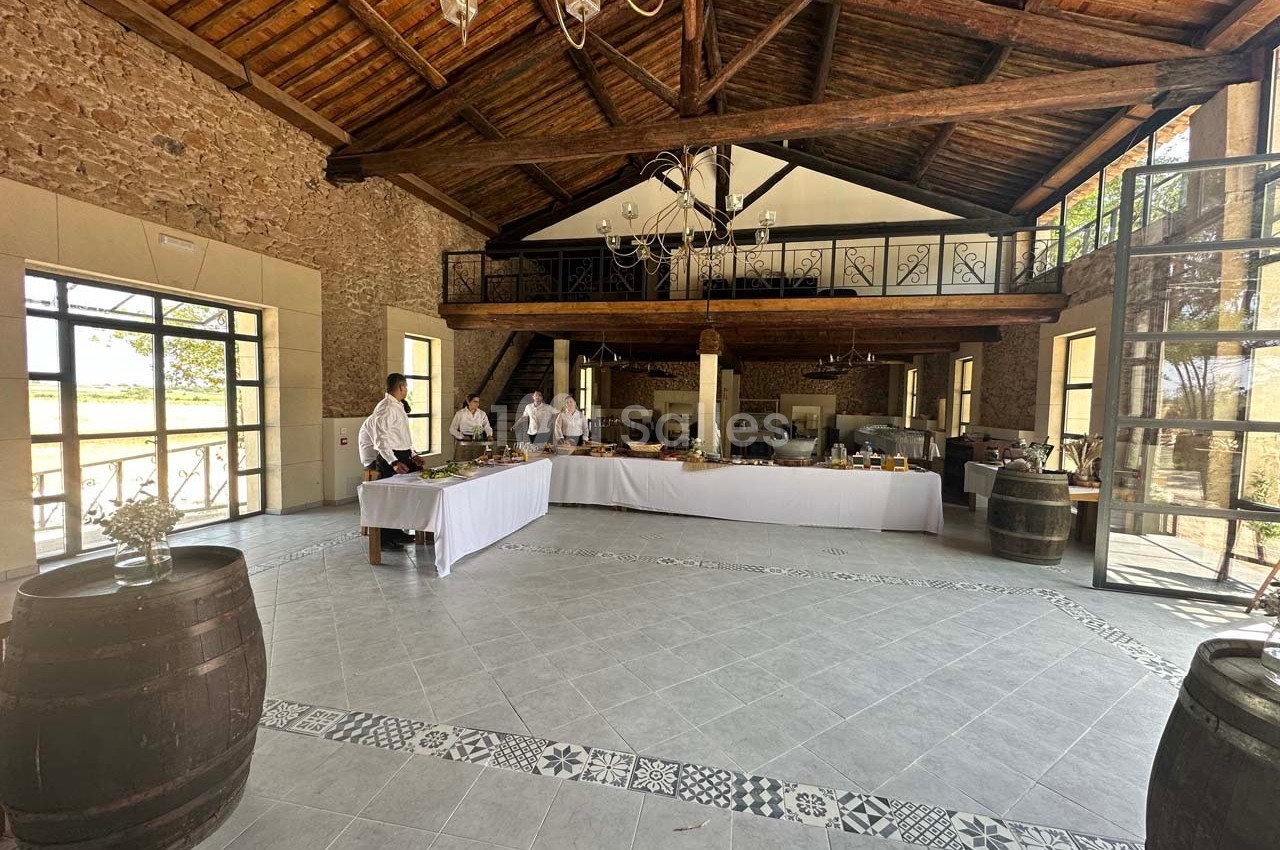 Salle spacieuse avec murs en pierre, plafond en bois, grandes fenêtres et tables dressées pour un buffet.