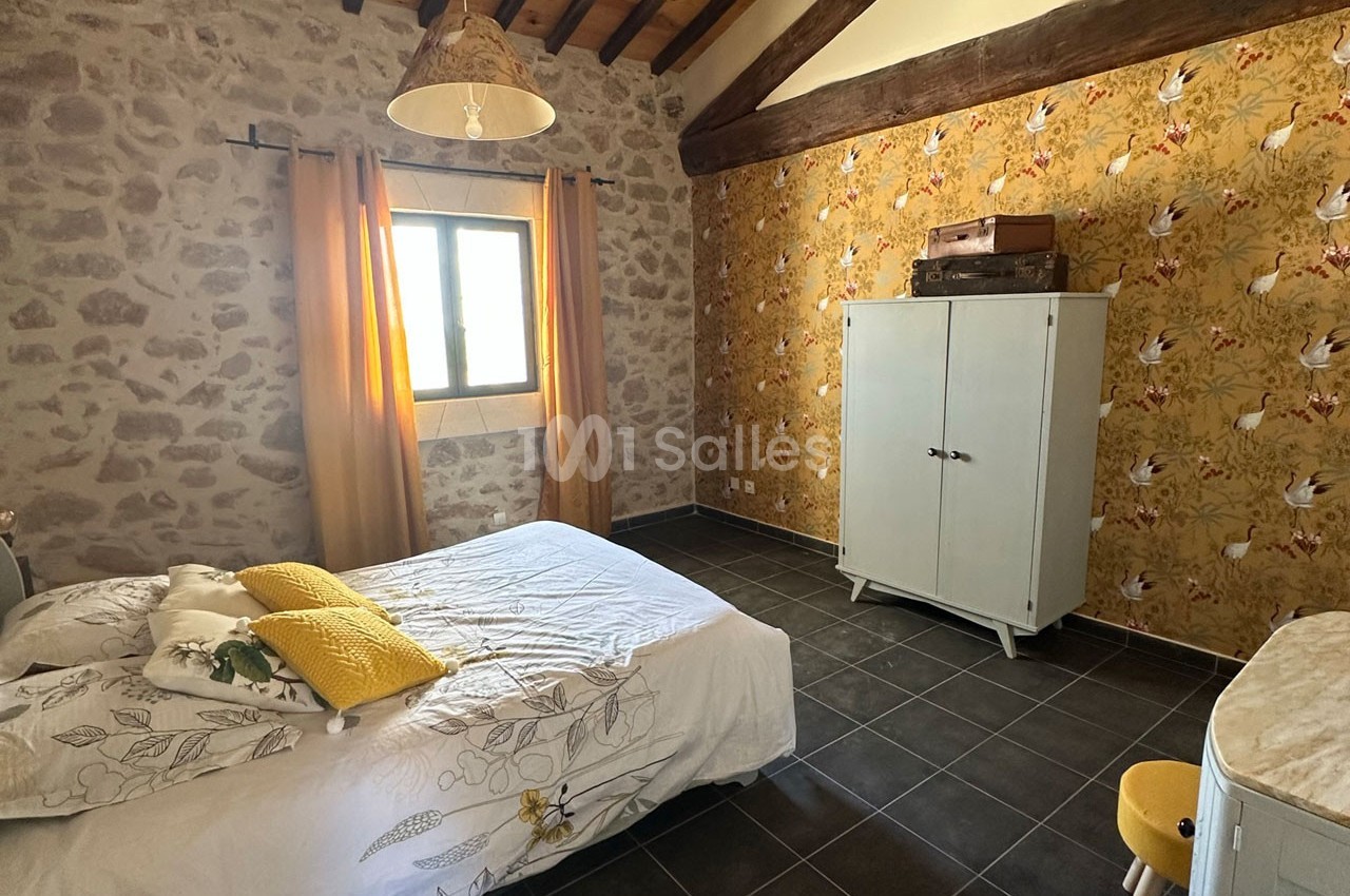 Chambre avec murs en pierre et papier peint floral, lit double, armoire blanche et fenêtre laissant entrer la lumière.