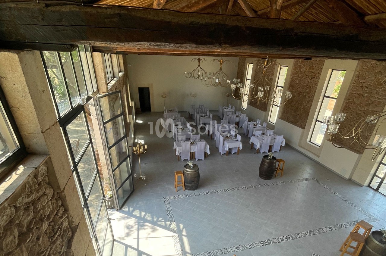 Salle de réception lumineuse avec tables dressées, murs en pierre, poutres apparentes et grandes fenêtres.