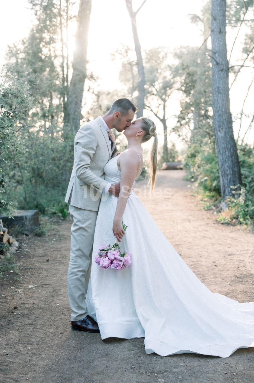 Un couple en tenue de mariage s'embrasse tendrement sur un chemin forestier, la mariée tenant un bouquet de fleurs roses.