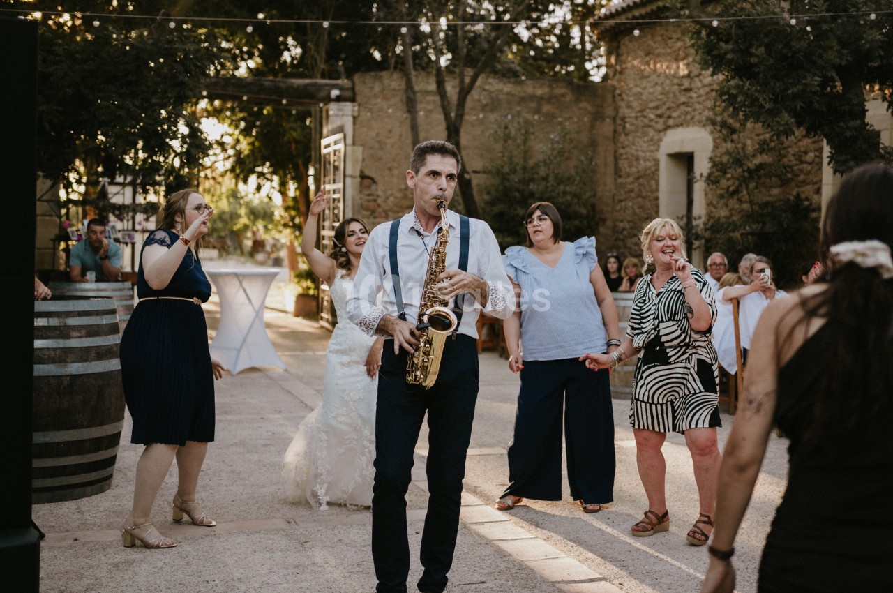 Un saxophoniste joue au milieu d'invités dans une ambiance festive en extérieur, près de bâtiments en pierre.