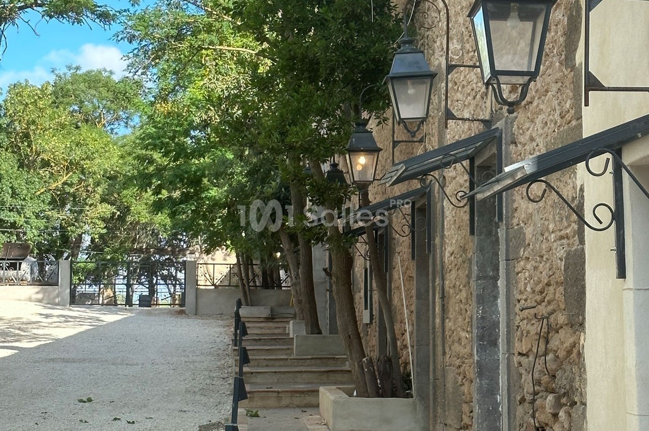 Allée bordée de lampadaires muraux et d'arbres, longeant un bâtiment en pierre sous un ciel dégagé.