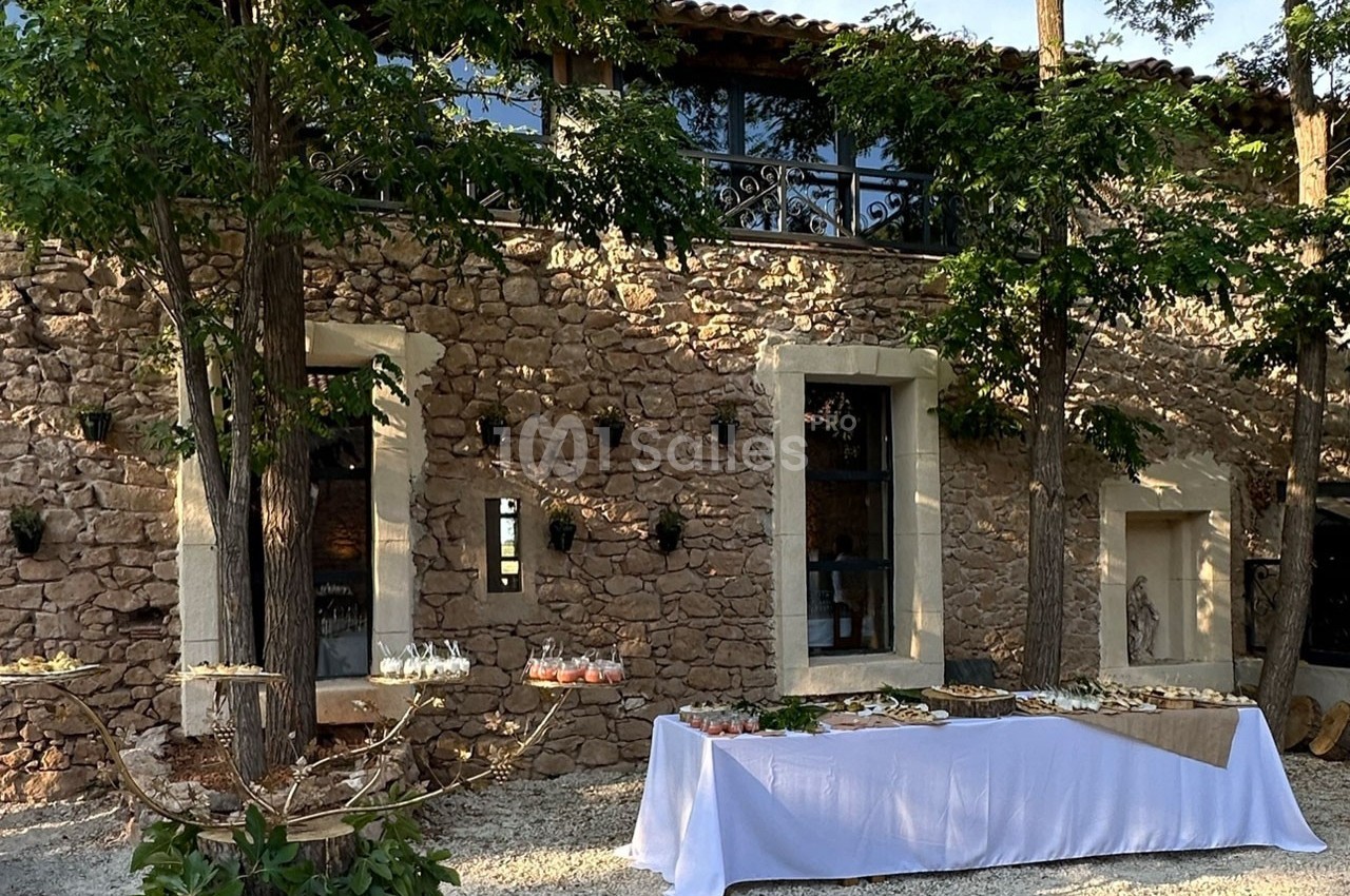Table dressée en extérieur devant une bâtisse en pierre, entourée d'arbres et de lumière naturelle.