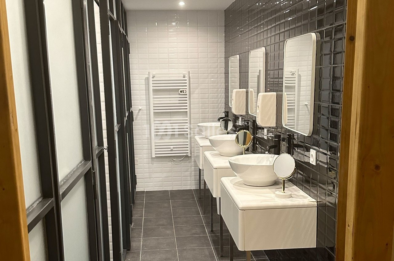 Salle de bain moderne avec trois lavabos, miroirs, carrelage noir et blanc, et un sèche-serviettes mural.