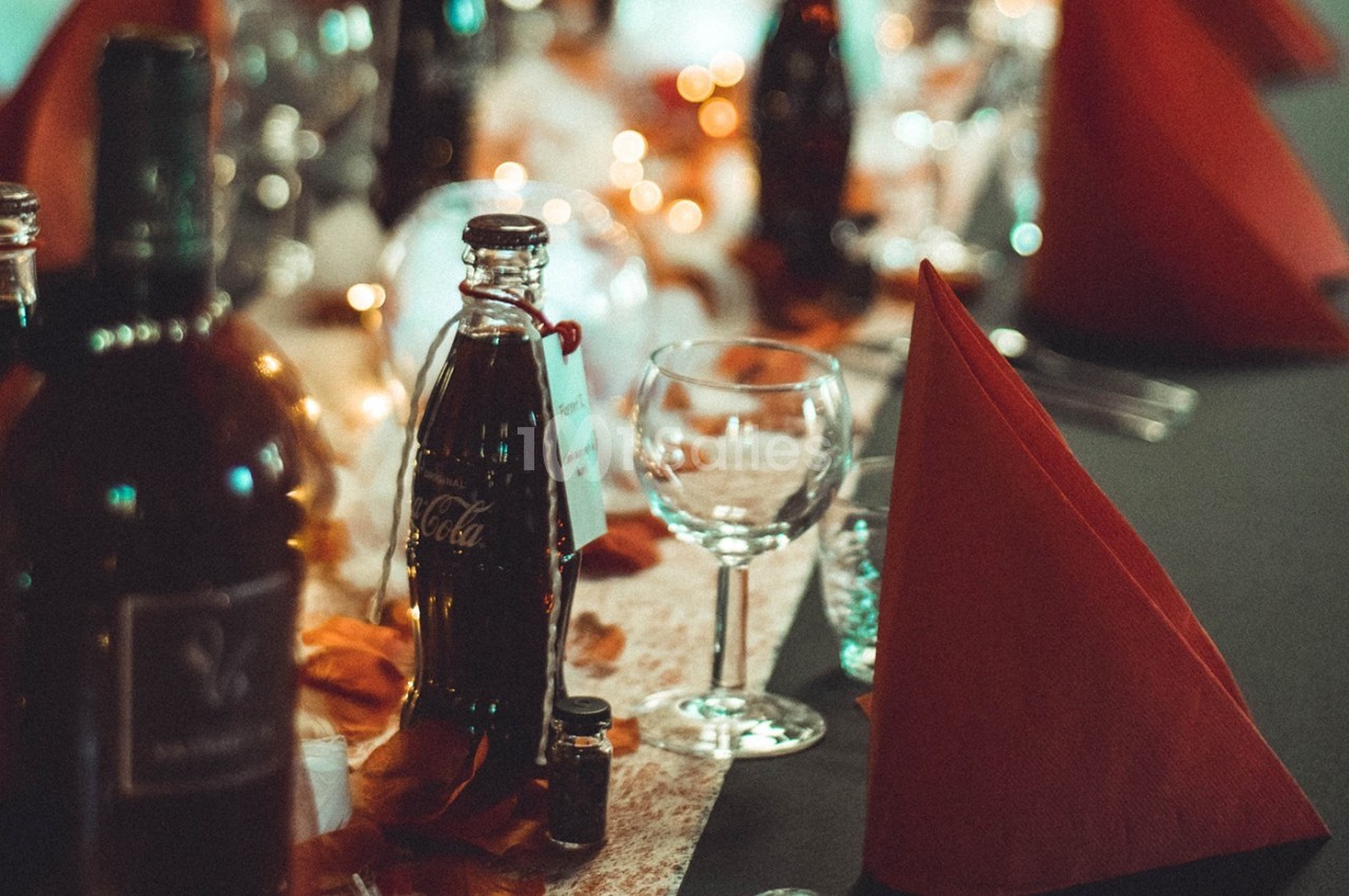 Table dressée avec des bouteilles, verres à pied, serviettes rouges pliées et décorations lumineuses.