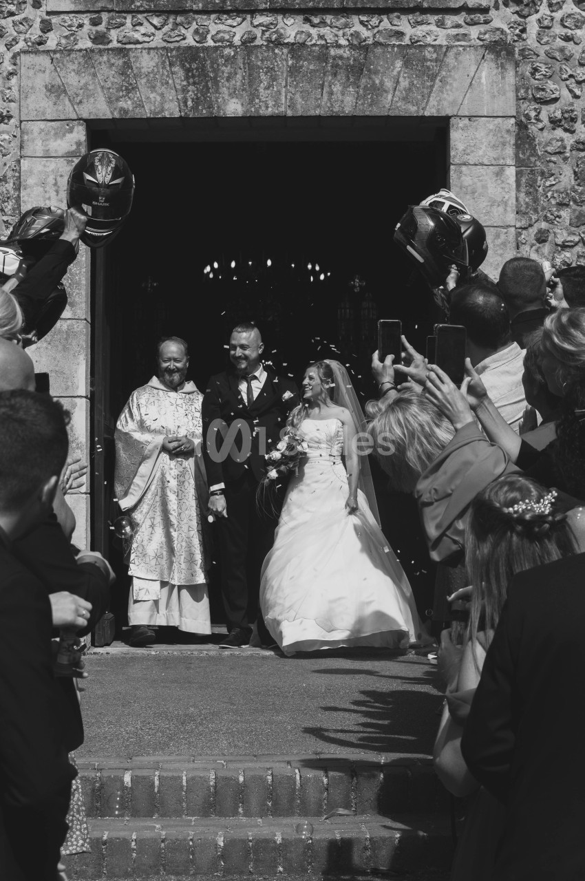Un couple de mariés sort d'une église, entouré d'invités tenant des ballons et prenant des photos.