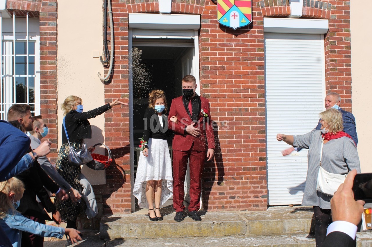 Un couple sort d'un bâtiment en tenue de mariage, entouré de personnes lançant des grains de riz.