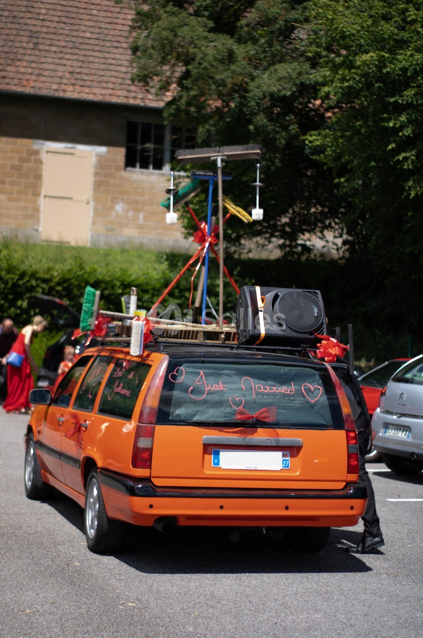 Voiture orange décorée pour un mariage avec des rubans, des accessoires et un message ’Just Married’ sur la vitre arrière.