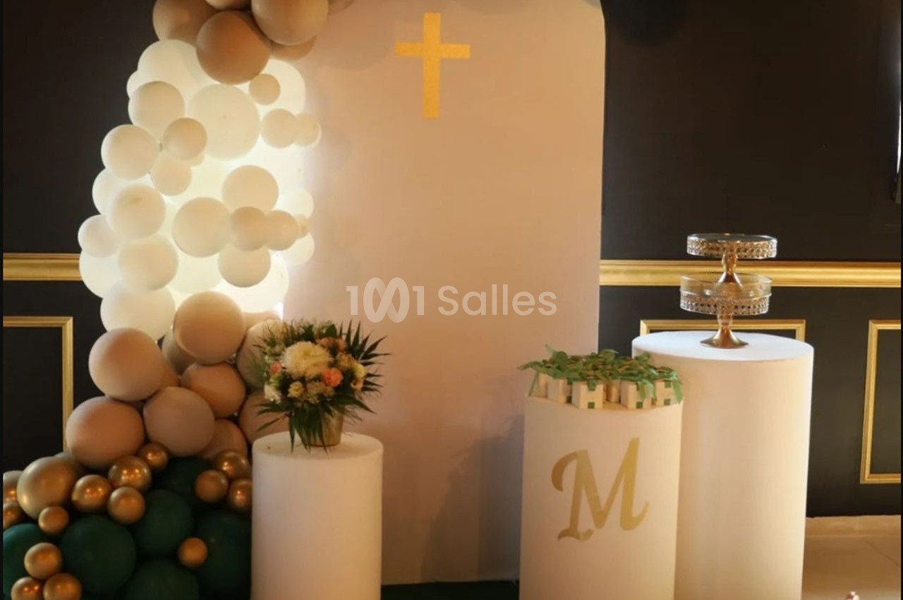 Décoration avec ballons, croix dorée, colonnes blanches, bouquet de fleurs et présentoir à gâteaux sur fond sombre.