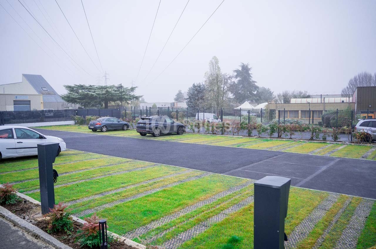 Parking extérieur avec des places alternant pavés et gazon, entouré de bâtiments et d'arbres par temps brumeux.