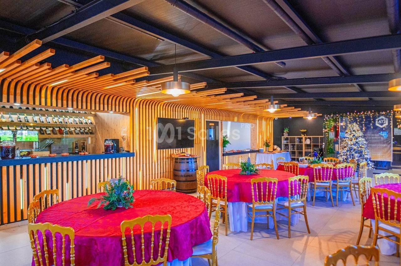 Salle de réception décorée avec des tables rondes, nappes rouges, chaises dorées et un bar en bois éclairé.