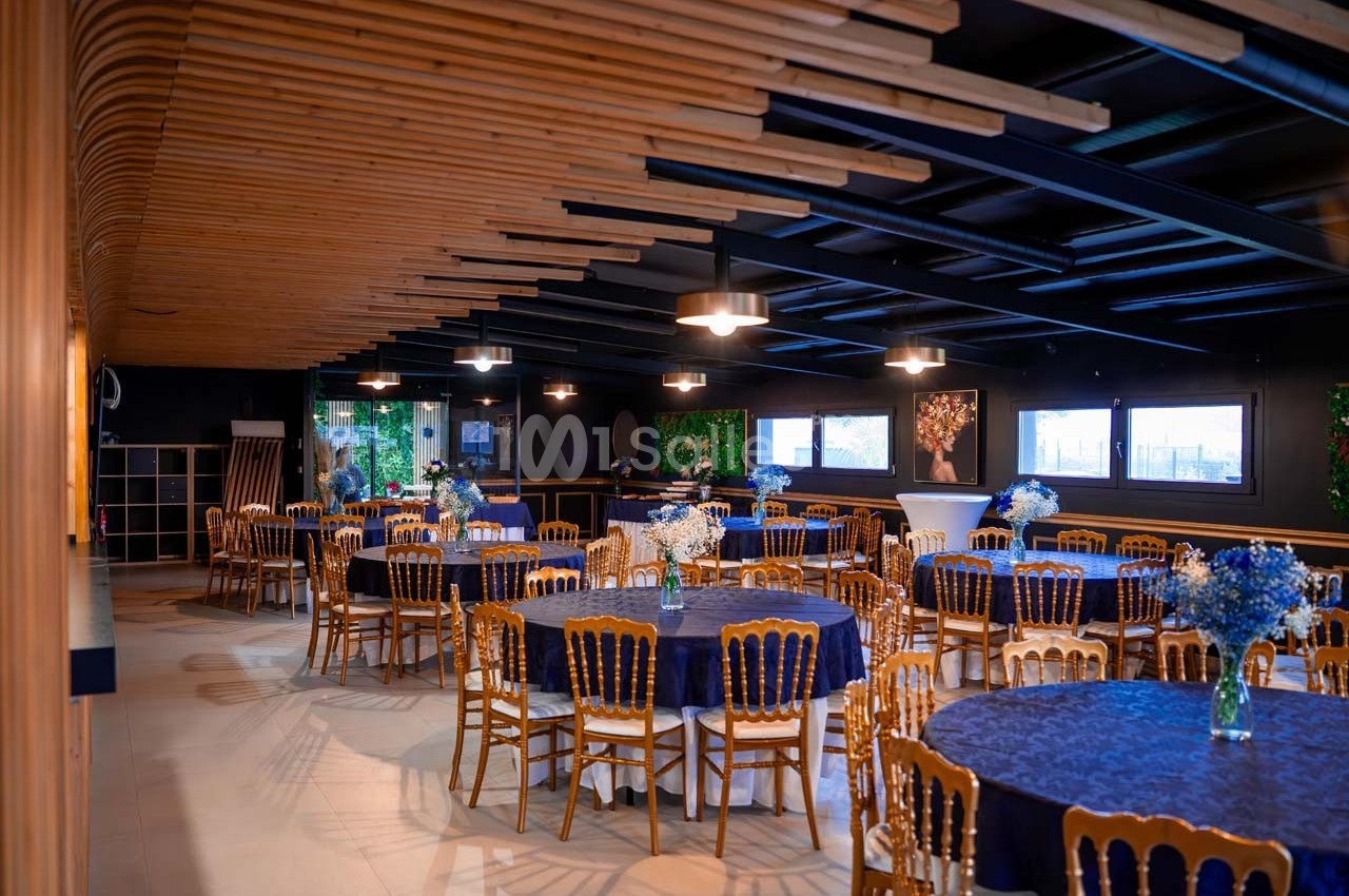 Salle de réception avec tables rondes dressées de nappes bleues, chaises en bois et décoration florale.