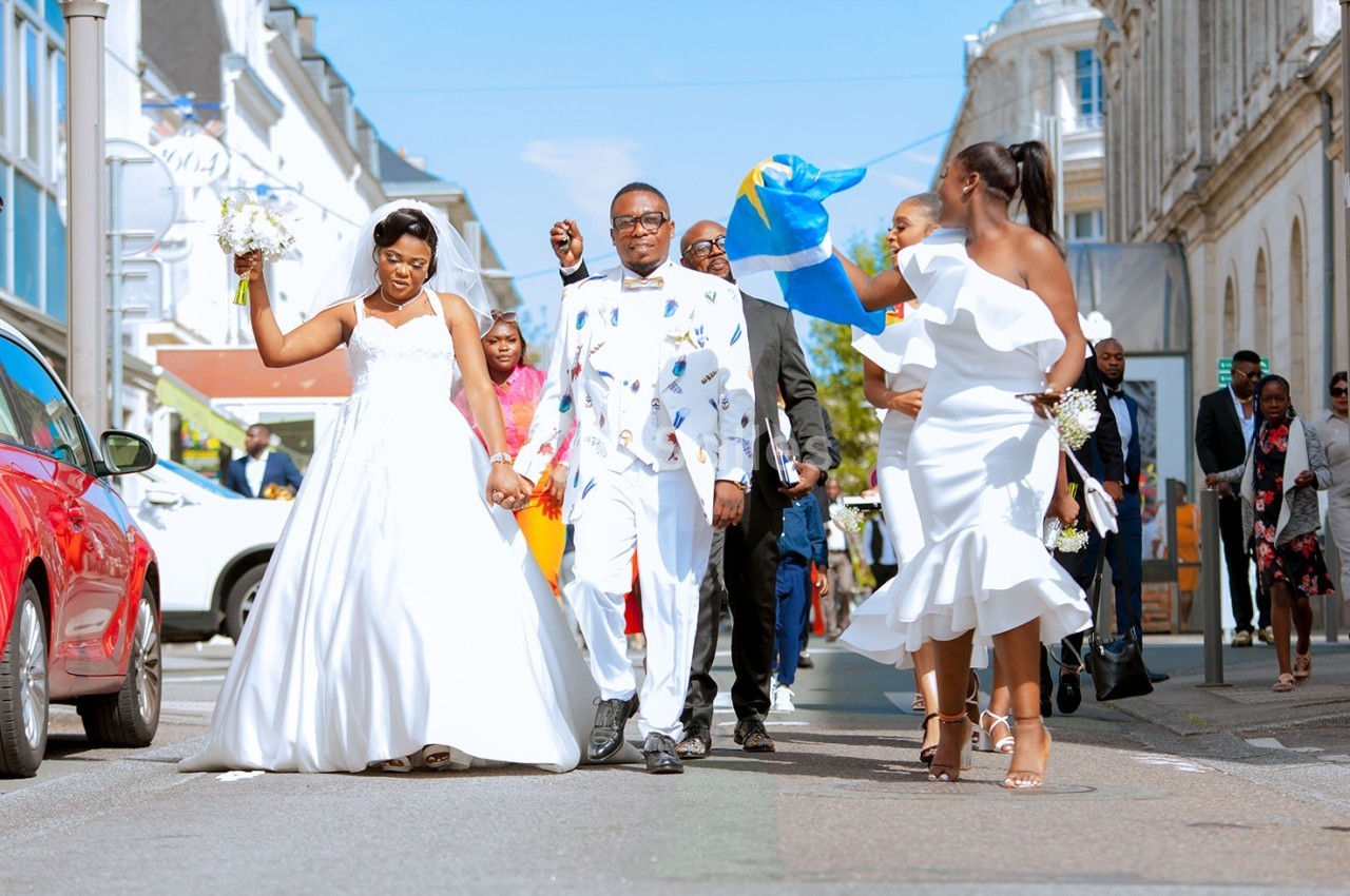 Un couple en tenue de mariage marche dans une rue, entouré d'invités festifs, dont l'un porte un drapeau.