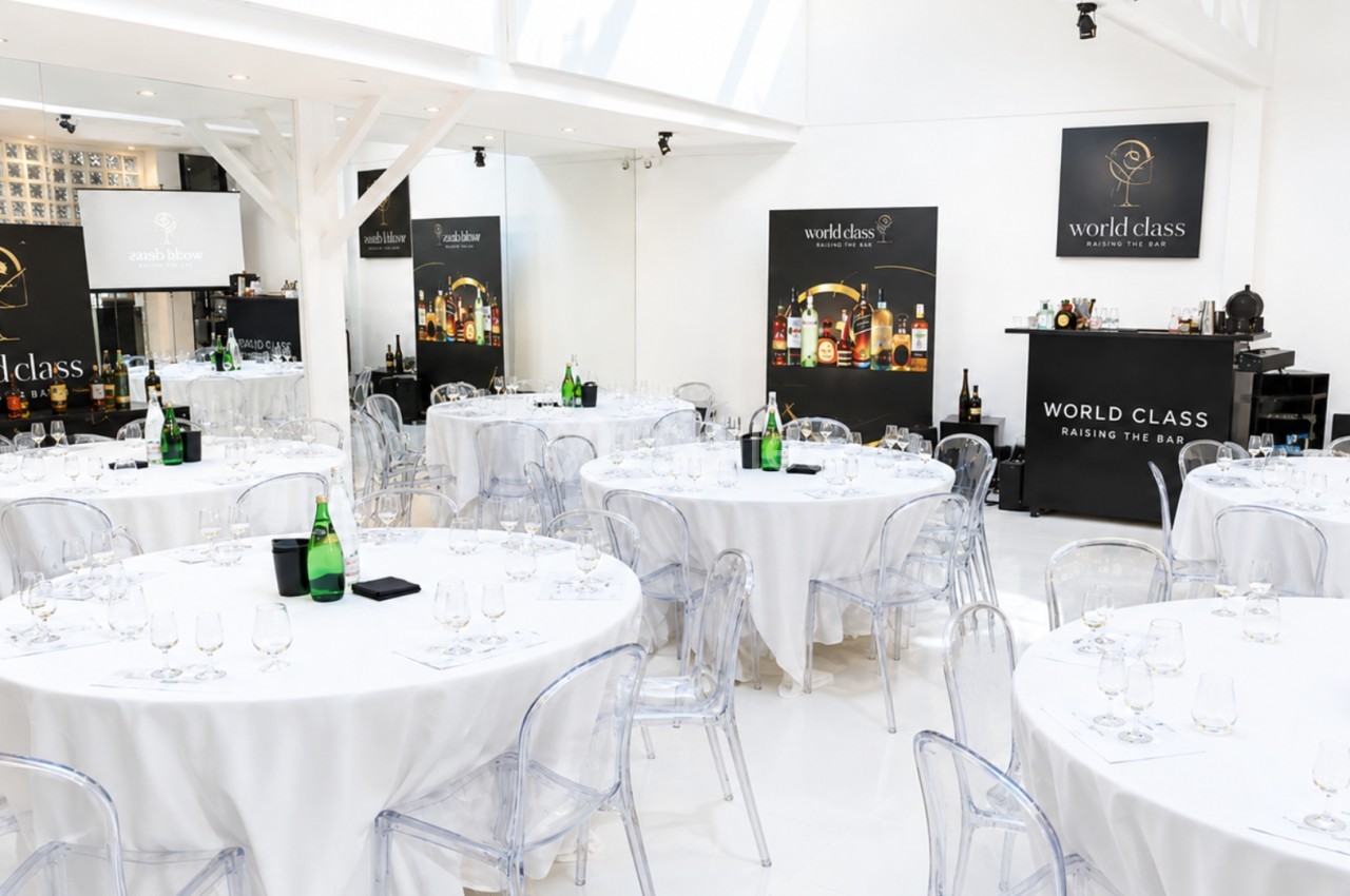 Salle lumineuse aménagée pour un événement, avec tables rondes dressées, chaises transparentes et bar en arrière-plan.