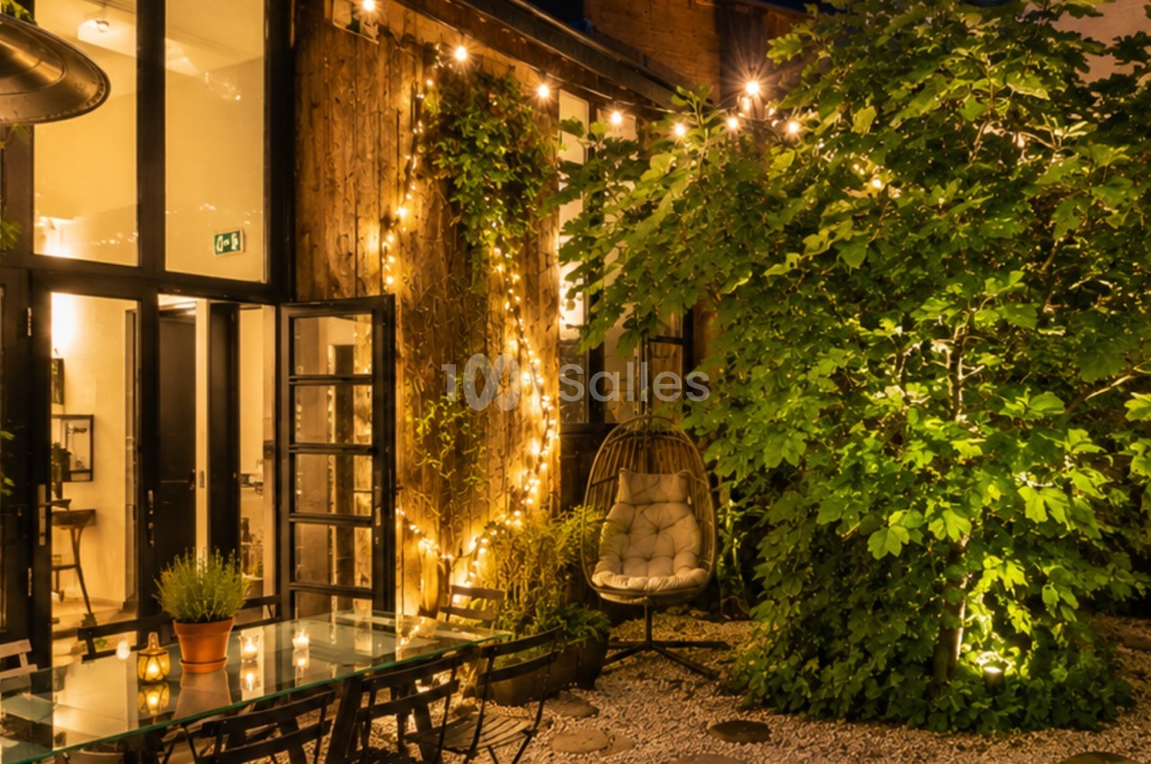 Terrasse éclairée par des guirlandes lumineuses, avec table en verre, chaises et végétation abondante en soirée.