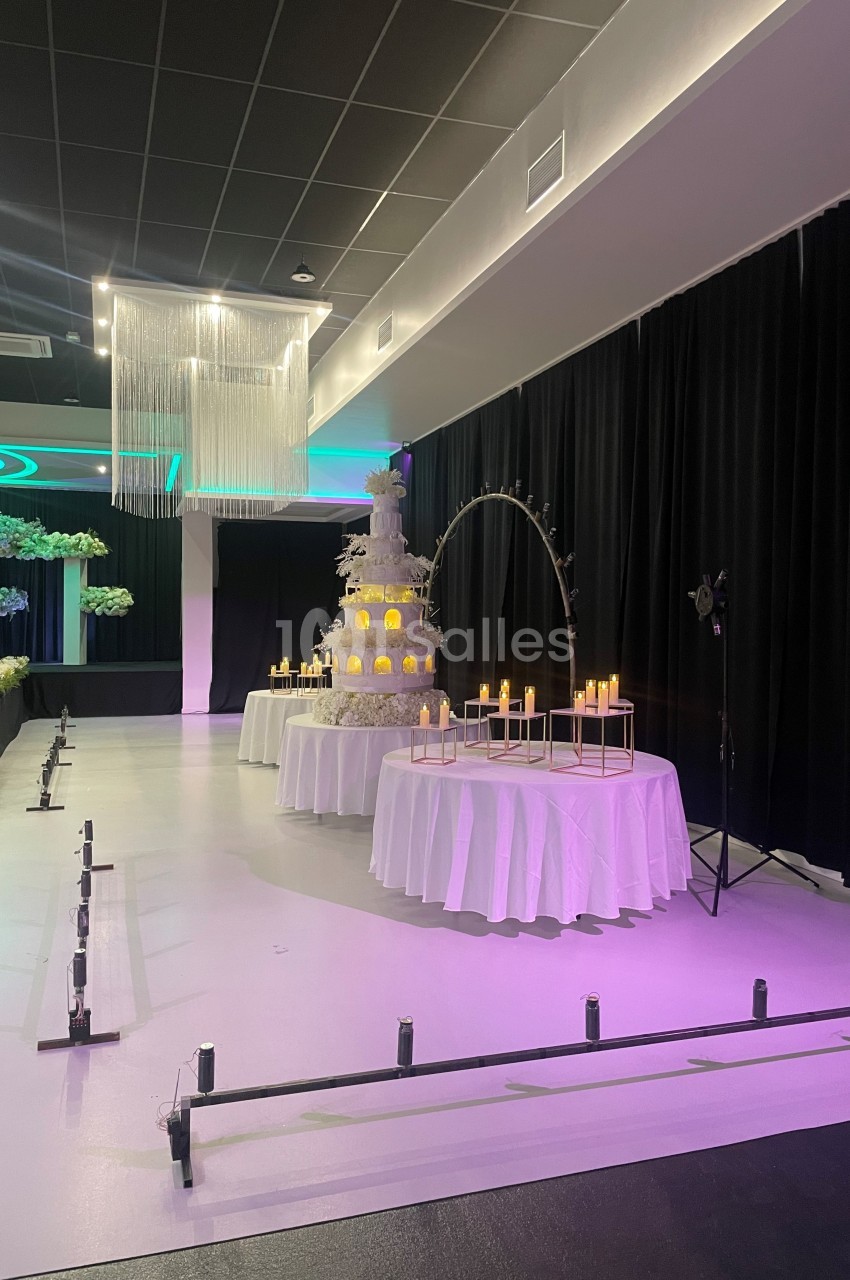Salle de réception décorée avec un grand gâteau de mariage éclairé, des bougies et des rideaux noirs en arrière-plan.