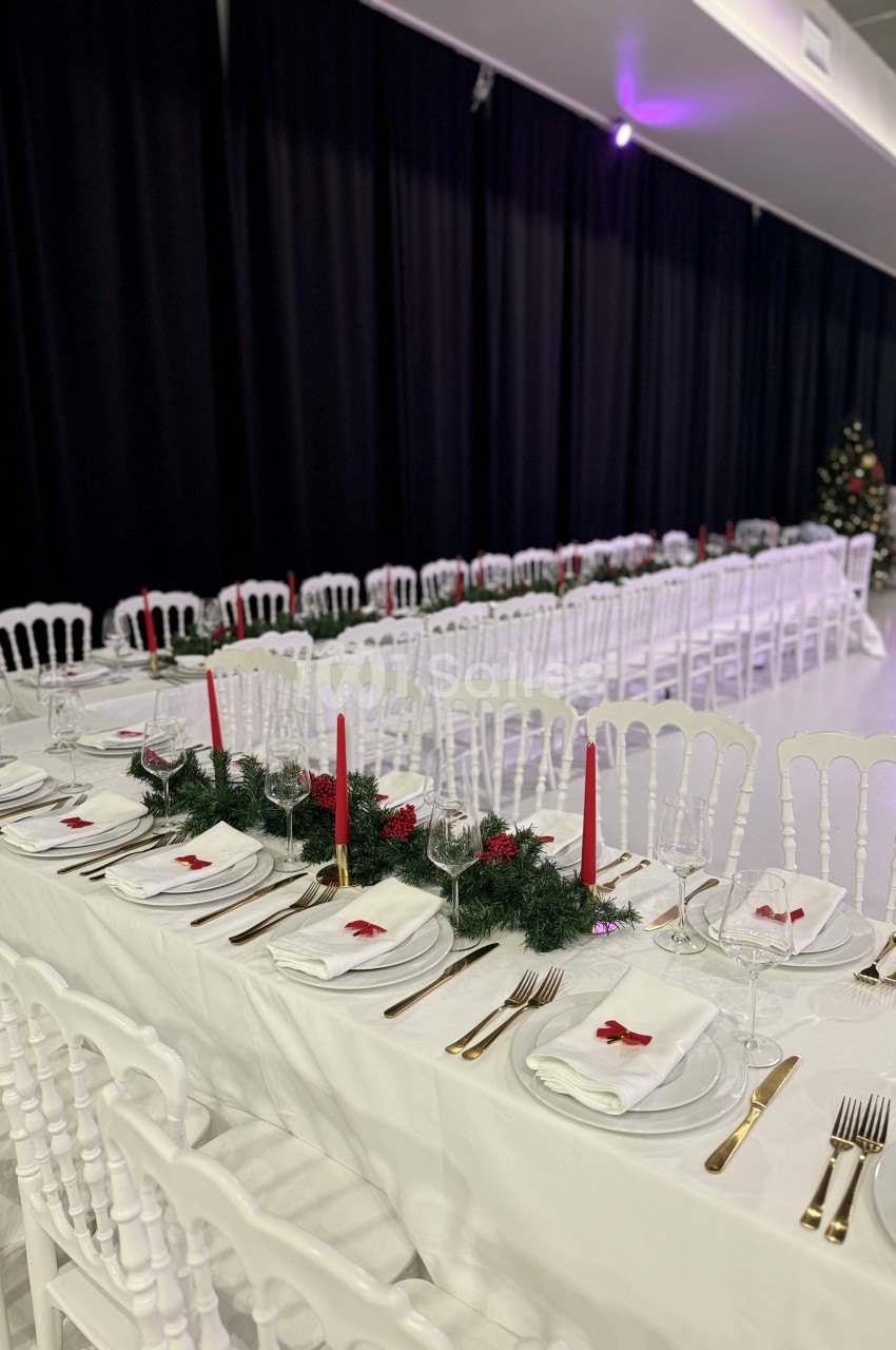 Tables de banquet décorées avec nappes blanches, bougies rouges, couverts dorés et guirlandes de sapin.