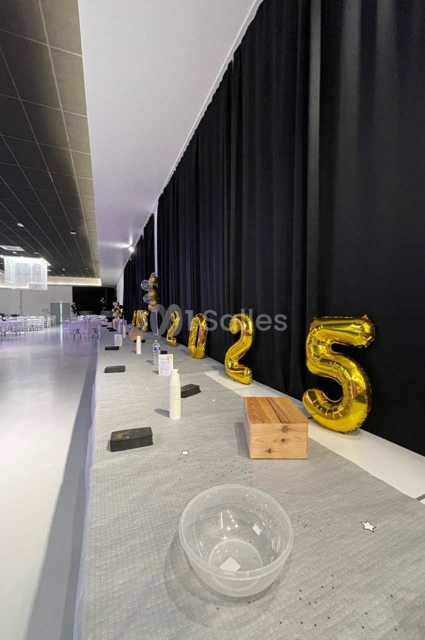 Ballons dorés formant ’2025’ alignés sur une table décorée, dans une salle avec rideaux noirs et chaises en arrière-plan.