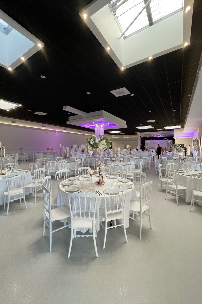 Salle de réception décorée avec des tables rondes dressées, chaises blanches et éclairage tamisé violet.