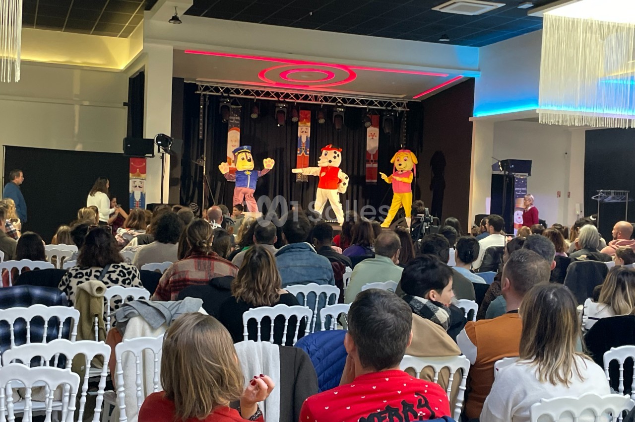 Des mascottes colorées dansent sur scène devant un public assis dans une salle de spectacle.