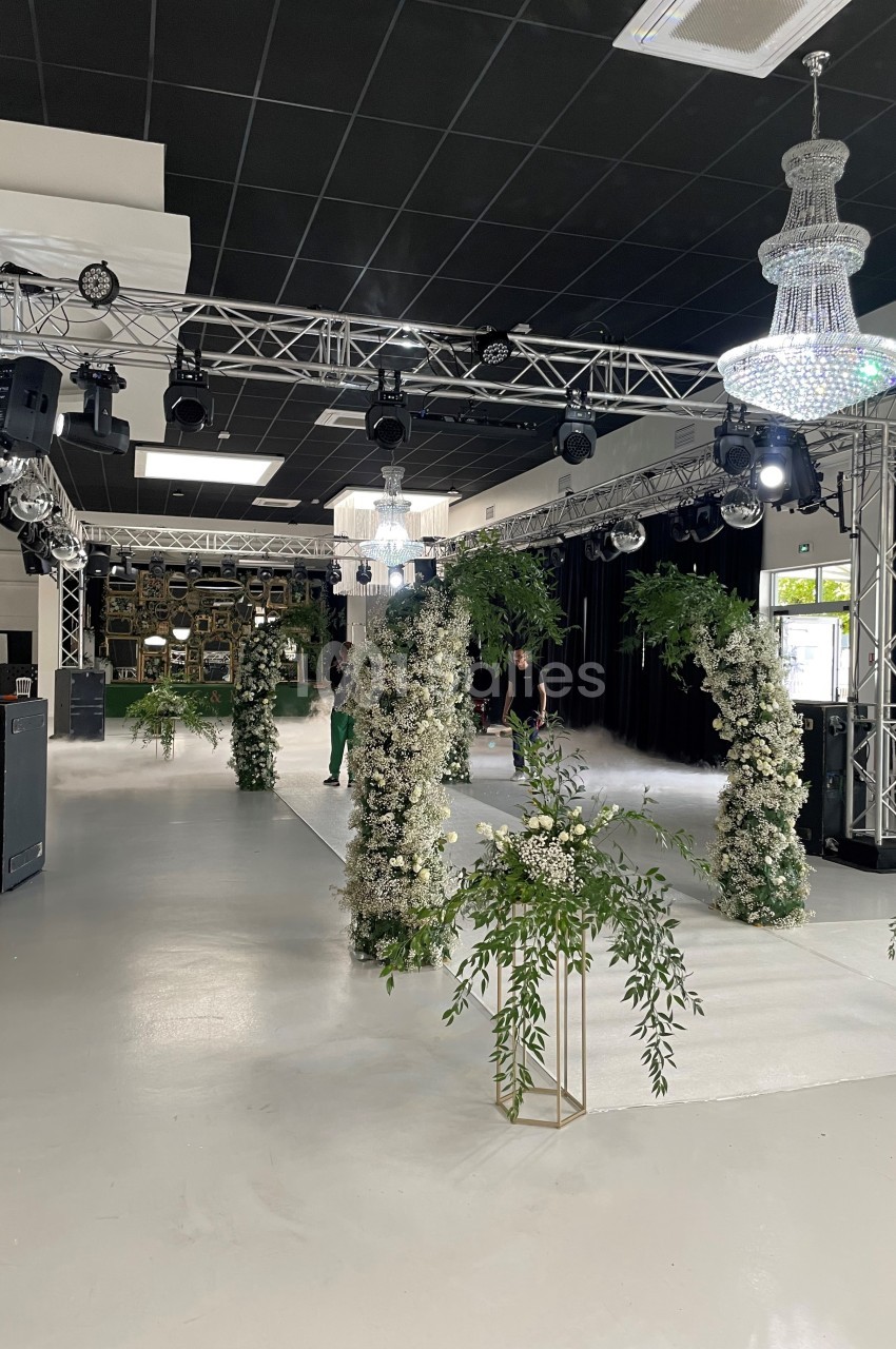Salle décorée pour un événement avec des arches fleuries, des lustres suspendus et une structure métallique éclairée.
