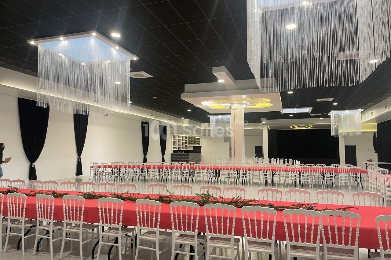 Salle de réception moderne avec tables rectangulaires dressées de nappes rouges, chaises blanches et décorations suspendues.