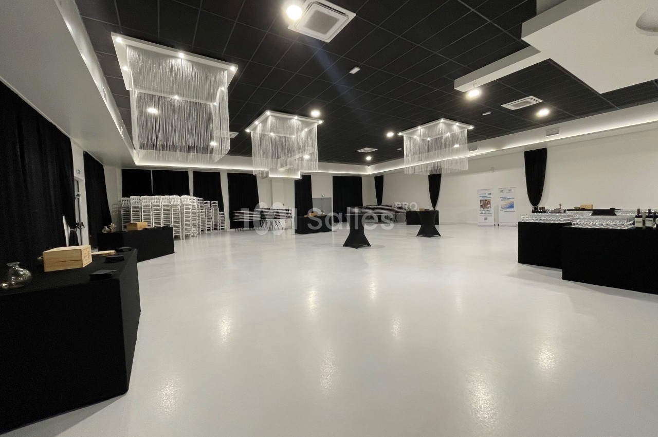 Salle spacieuse avec sol clair, tables hautes noires, chaises empilées et éclairage moderne suspendu.