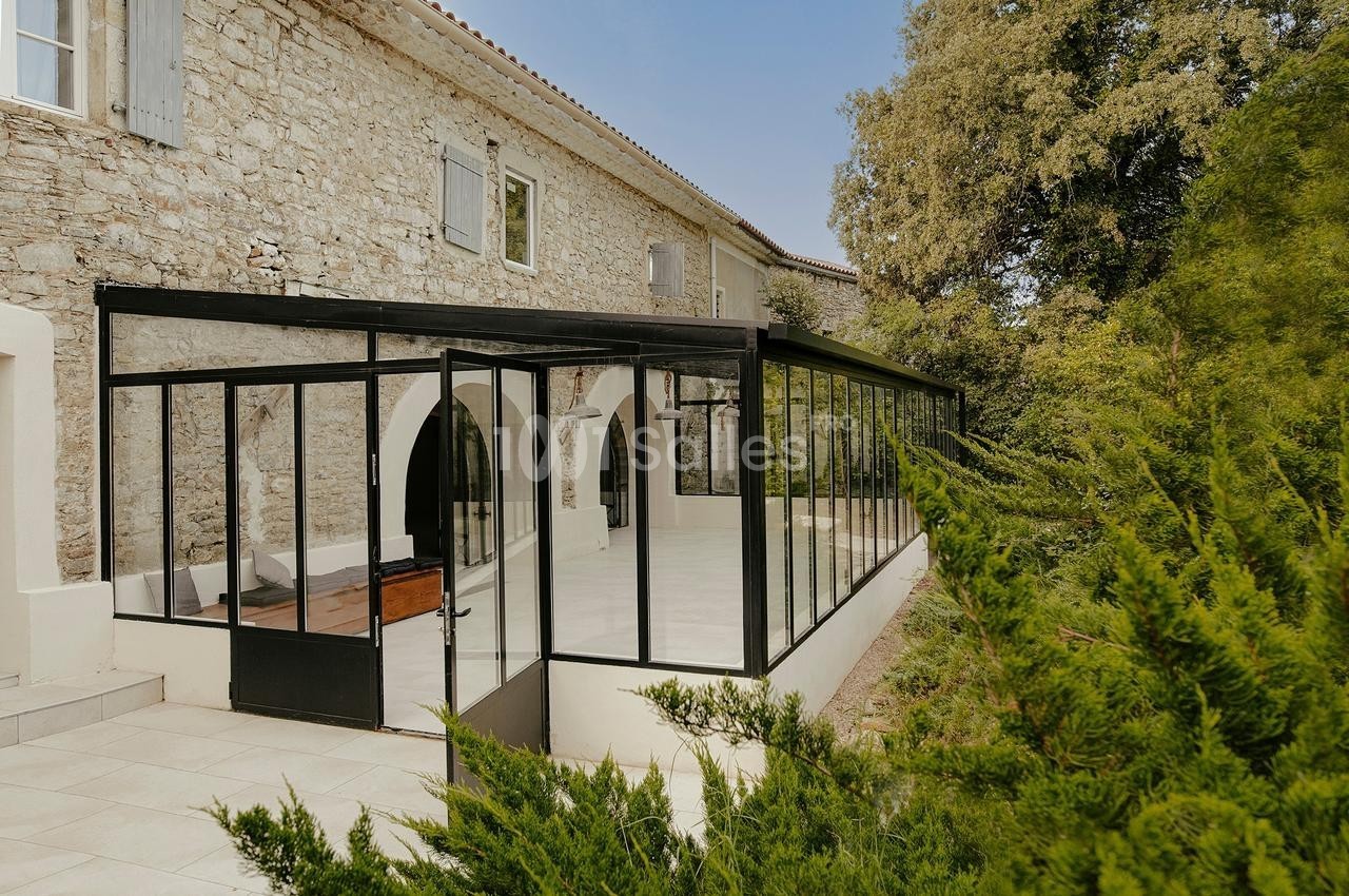 Véranda moderne en structure noire attenante à une maison en pierre, entourée de végétation.