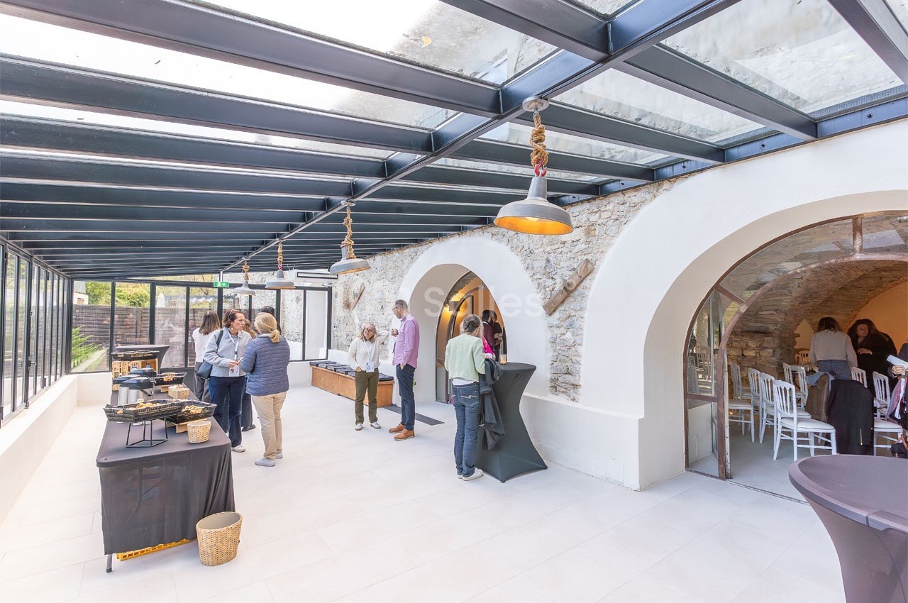 Salle lumineuse avec verrière, murs en pierre, tables de buffet et personnes discutant ou se servant.