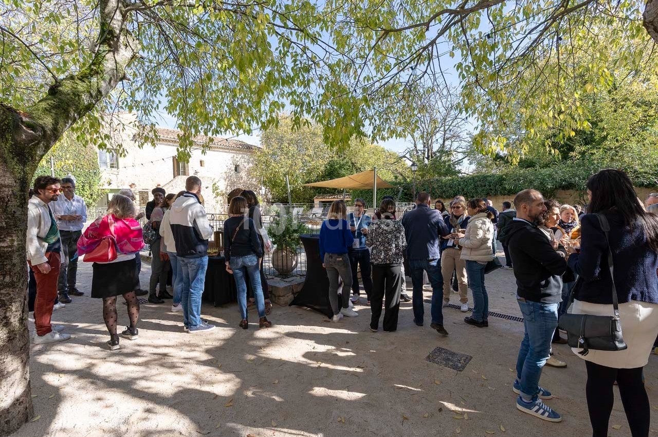 Groupe de personnes discutant en extérieur sous des arbres, près de tables et d'un bâtiment en pierre.