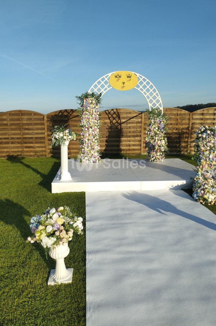 Arche de mariage décorée de fleurs pastel sur une estrade blanche, entourée de pelouse et de ciel dégagé.