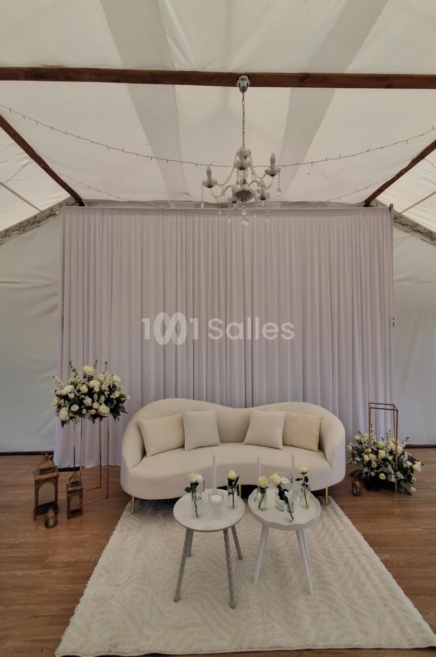 Canapé beige avec coussins, tables basses décorées de fleurs, tapis blanc, rideau et lustre dans une tente élégante.
