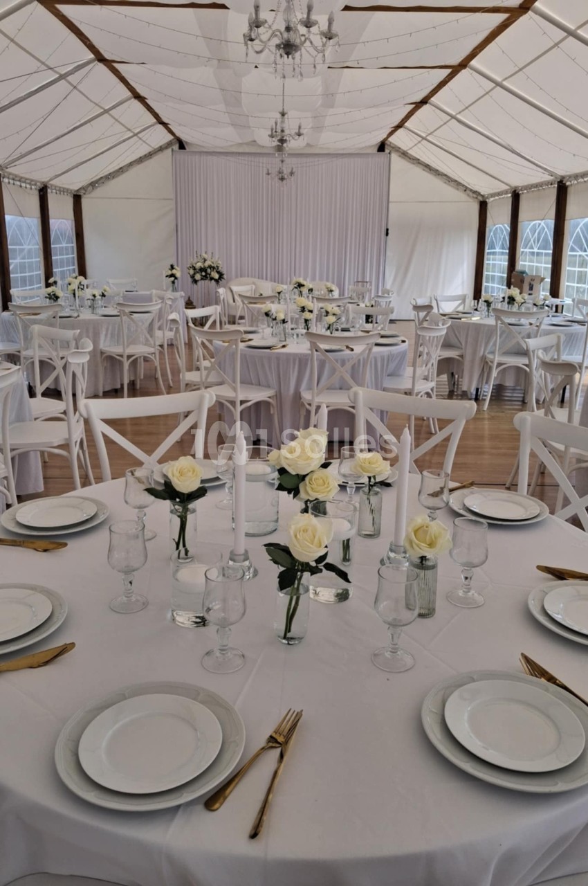 Salle de réception décorée avec des tables rondes dressées, nappes blanches, roses blanches et couverts dorés.