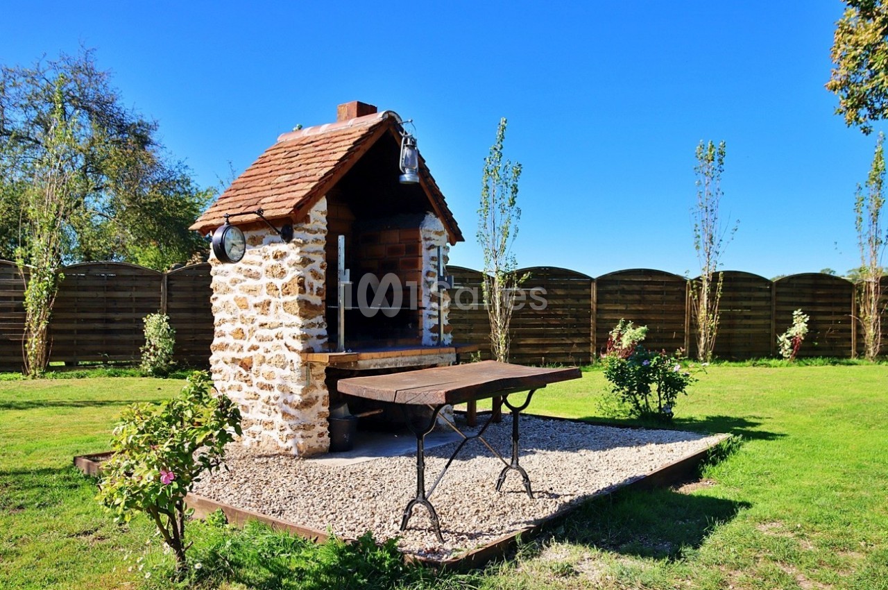 Barbecue en pierre avec toit en tuiles, table en bois et gravier environnant, situé dans un jardin ensoleillé.