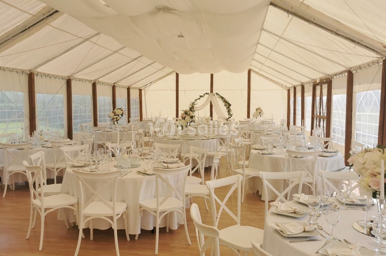 Salle de réception décorée avec des tables rondes, nappes blanches, chaises en bois et arche fleurie au fond.