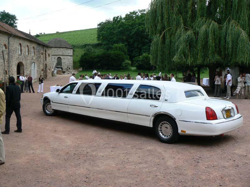 Une limousine blanche garée sur un chemin en gravier, avec des invités rassemblés près d'un bâtiment en pierre.