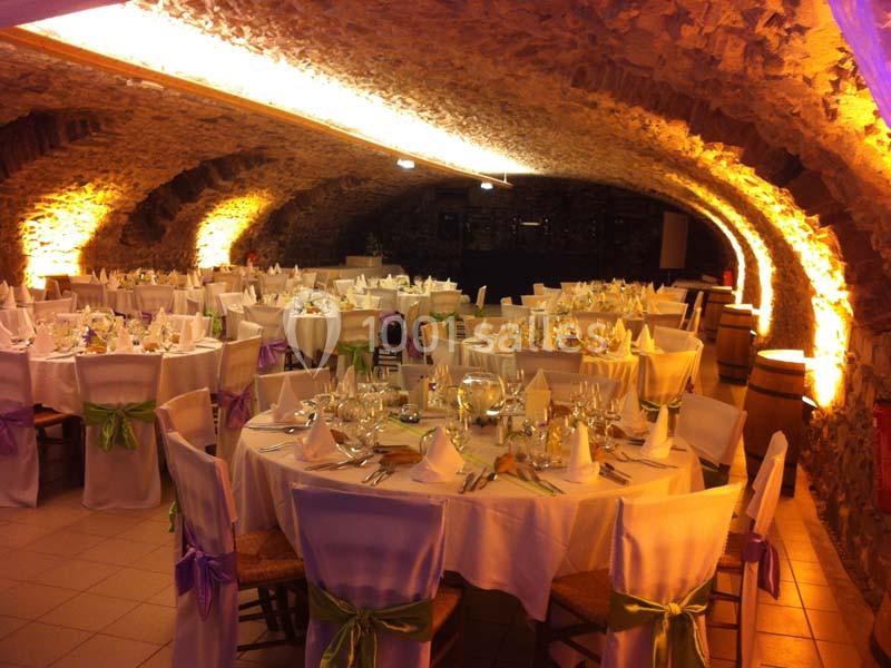 Salle voûtée en pierre aménagée pour un banquet, avec tables rondes dressées et décorations colorées.
