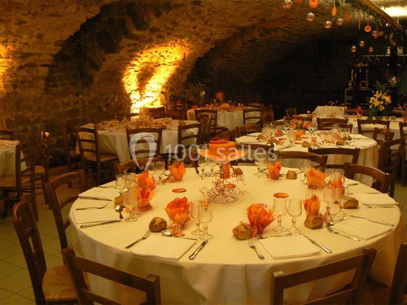 Salle voûtée en pierre avec tables rondes dressées pour un repas, décorées de serviettes et centres de table orange.
