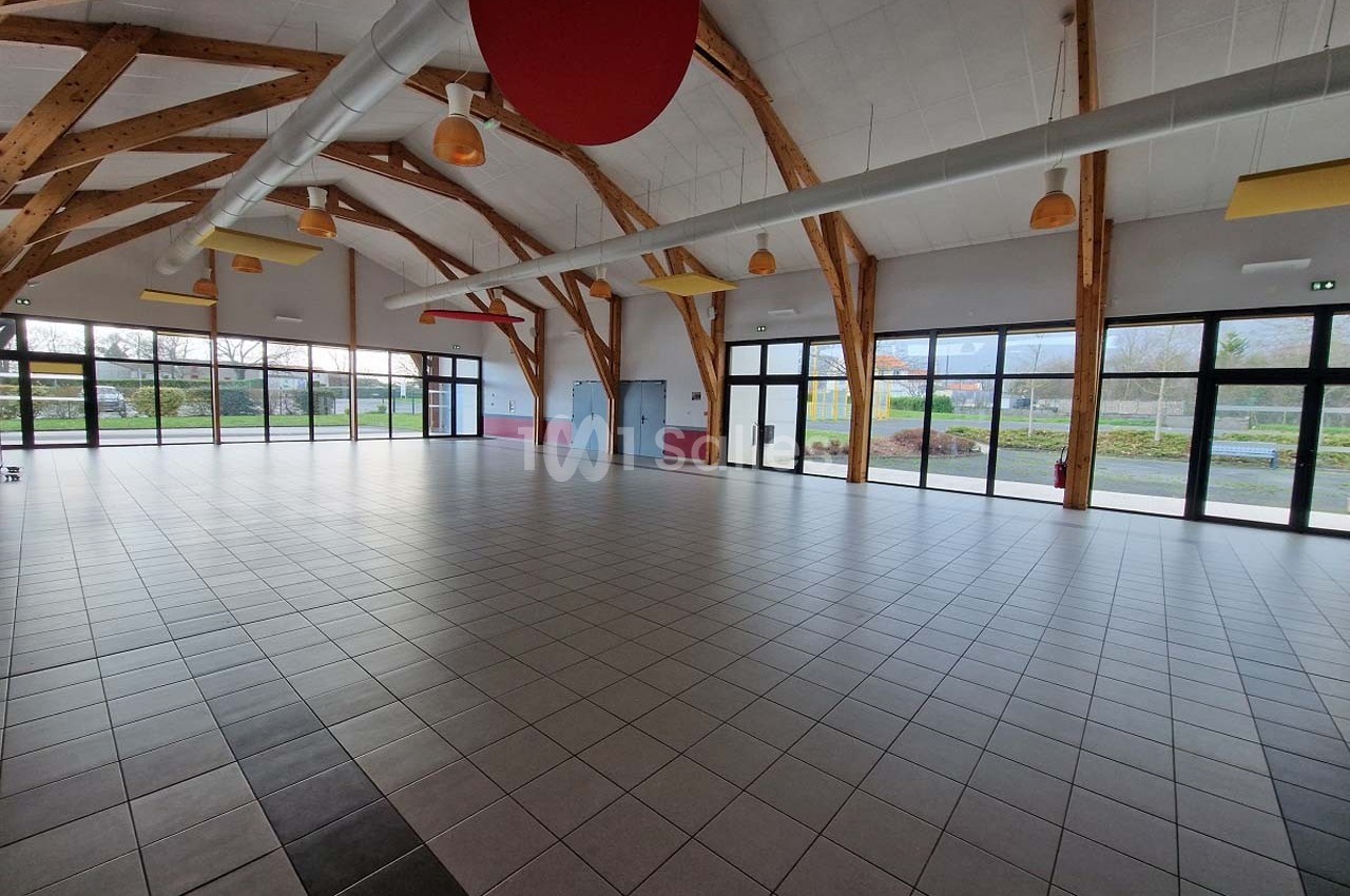 Grande salle avec charpente en bois apparente, sol carrelé et larges baies vitrées donnant sur un espace extérieur.