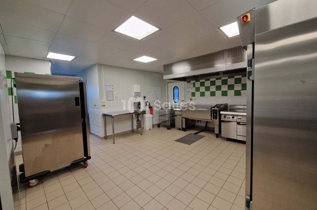 Cuisine professionnelle avec équipements en inox, plan de travail, four, réfrigérateur et carrelage au sol beige.