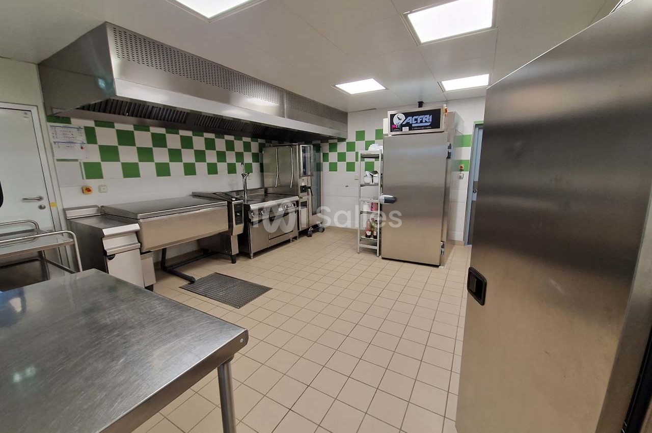 Cuisine professionnelle avec équipements en inox, carrelage beige au sol et murs partiellement carrelés en blanc et vert.