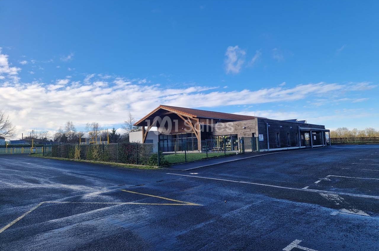 Bâtiment moderne en bois avec un toit en pente, entouré d'une clôture et situé près d'un parking asphalté.