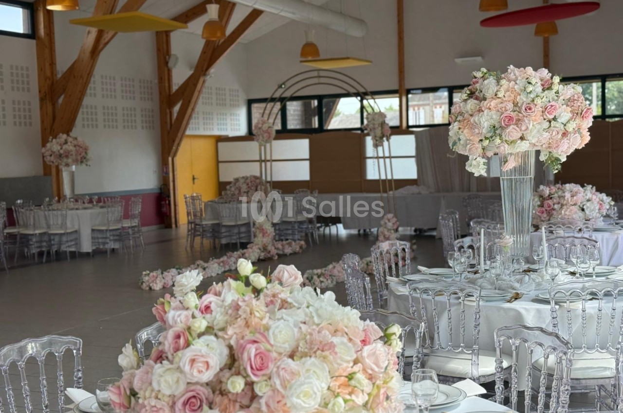 Salle de réception décorée avec des tables rondes, des chaises transparentes et des compositions florales roses et blanches.