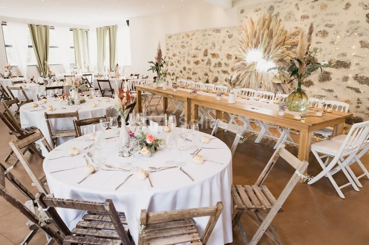 Salle de réception décorée avec des tables rondes et rectangulaires, chaises en bois, fleurs et lumière naturelle.